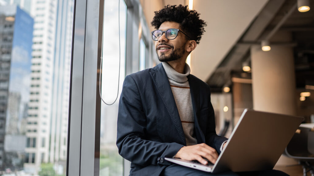 A man sitting beside window and looking outside while working on laptop