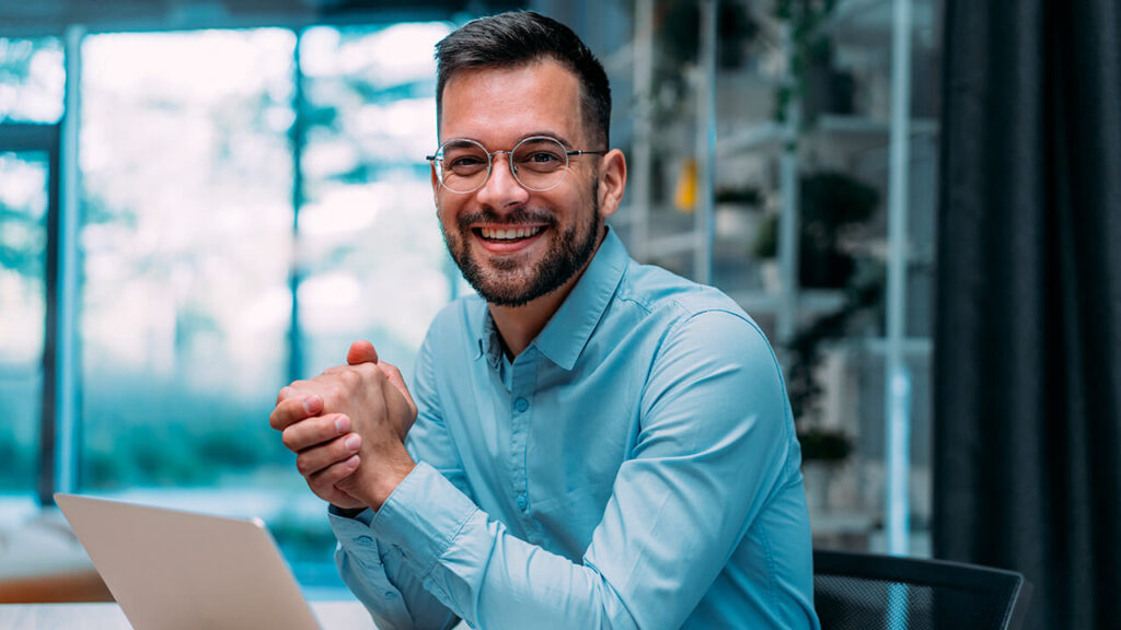 A man wearing a blue button up collard shirt with his hands clasped smiling.